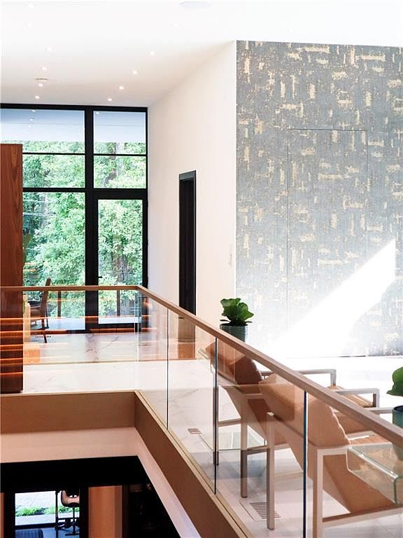 This modern interior view captures a mezzanine landing overlooking a lower level, featuring a glass railing and light-toned flooring. A prominent wall with a textured, metallic-patterned finish serves as a focal point, while large windows provide an expansive view of the surrounding greenery. The perspective emphasizes the open, airy architectural design and the seamless transition between indoor living spaces.