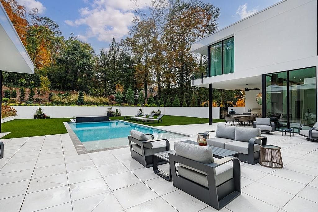 This modern, minimalist residence features a spacious outdoor living area centered around a rectangular swimming pool with a dark-tiled water feature. The patio is paved with large, light-colored tiles and furnished with contemporary grey lounge seating, while a manicured lawn and mature trees provide a lush backdrop. The home's architecture is defined by clean white lines and expansive glass walls that seamlessly connect the indoor and outdoor spaces.
