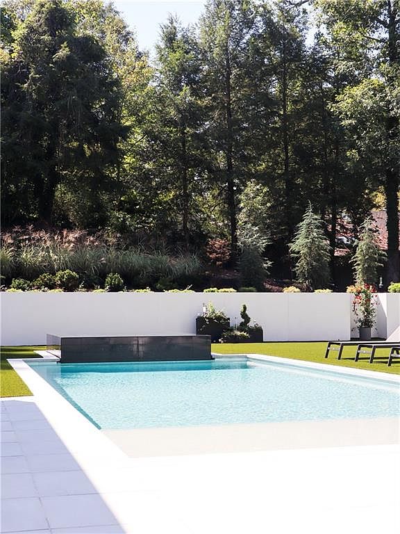 This image captures a modern, minimalist swimming pool area set against a backdrop of lush, mature trees. The pool features a sleek, dark-toned water feature at one end, contrasting with the bright white perimeter wall and clean-lined patio decking. The scene is framed from a low, eye-level perspective, emphasizing the serene and private atmosphere of the outdoor living space.