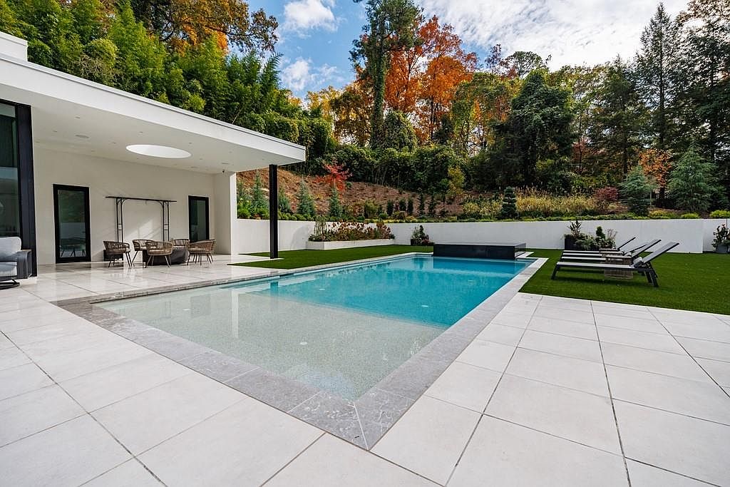 This modern outdoor living space features a rectangular swimming pool with a dark-tiled spa edge, set against a backdrop of lush, autumn-colored trees. A covered patio area with light-colored stone tiling provides a shaded lounge space, while a manicured lawn area hosts several sleek lounge chairs. The clean lines of the white architectural structure and the surrounding stone walls create a sophisticated, resort-like atmosphere.