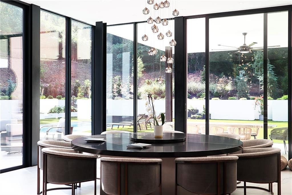 This sophisticated dining room features a large, circular dark-toned table surrounded by modern, upholstered chairs with dark frames. The space is defined by expansive floor-to-ceiling windows that provide a seamless connection to the outdoor pool and garden area. A striking, multi-tiered crystal pendant light fixture hangs above the table, serving as a focal point in this bright, contemporary interior.