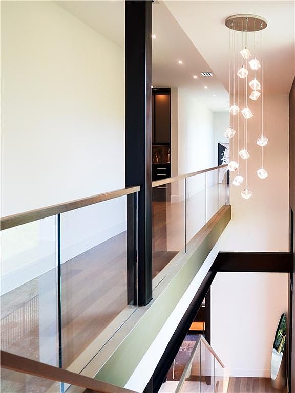 This perspective captures a modern, open-concept landing area overlooking a staircase, defined by a sleek glass railing with a dark metal frame. A striking, multi-tiered crystal pendant light fixture hangs from the ceiling, casting a warm glow over the space. The clean lines and minimalist aesthetic create an airy, sophisticated atmosphere that connects the upper level to the lower floor.