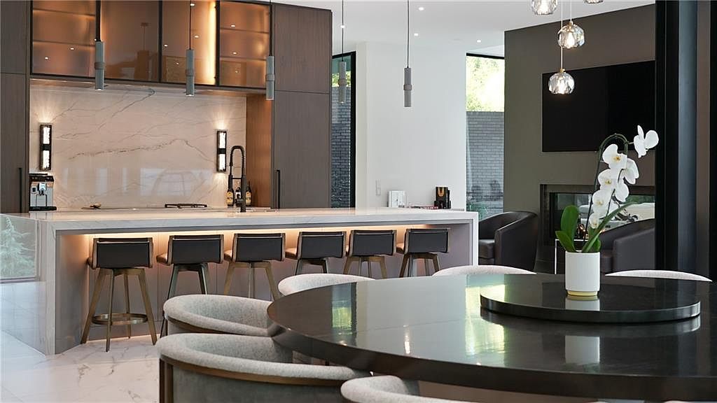 This sophisticated, modern kitchen features a large island with bar seating and a stone countertop, serving as the focal point of the open-concept space. Dark wood cabinetry contrasts with a light stone backsplash, while sleek pendant lights hang above the island and a dining area. The perspective captures the seamless flow between the kitchen and an adjacent living area, which includes a dark-toned wall with a built-in television and fireplace.