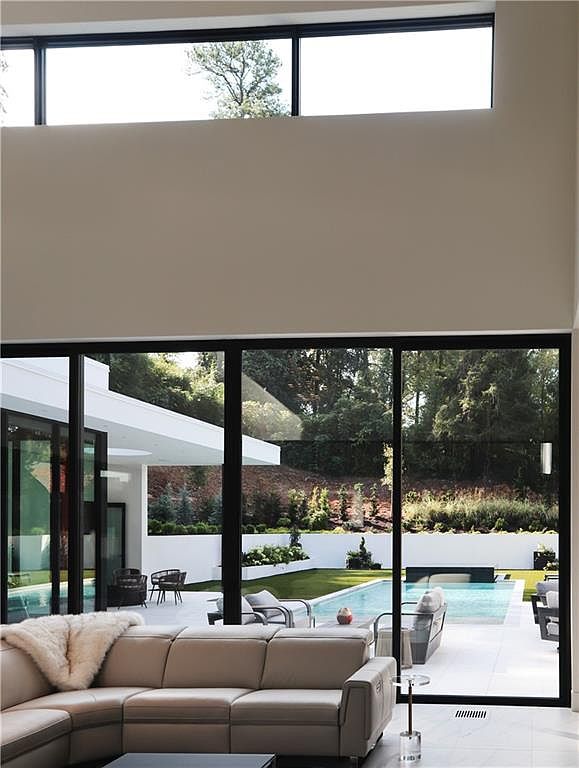This modern living room features a neutral-toned sectional sofa and expansive glass sliding doors that provide a seamless transition to the outdoor patio and pool area. The space is characterized by high ceilings with a clerestory window that enhances the natural light, creating an airy and sophisticated atmosphere. The view through the glass highlights a manicured backyard with a swimming pool, emphasizing a luxurious indoor-outdoor lifestyle.