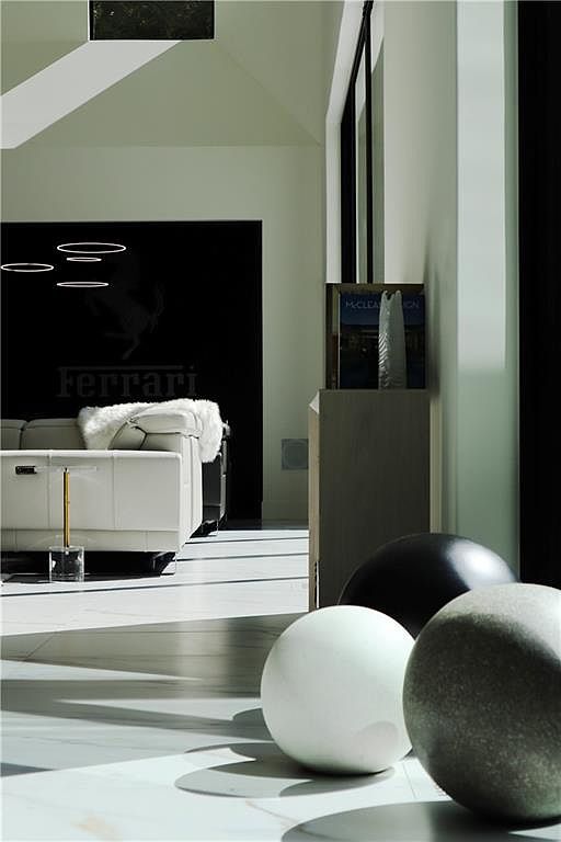 This modern living space features a sleek white leather sofa positioned against a striking black wall adorned with a Ferrari logo. The room is illuminated by contemporary ring-shaped pendant lights and natural light streaming in from the right, casting long shadows across the light-colored stone flooring. In the foreground, three large, spherical decorative objects in varying shades of white, black, and grey add a sculptural element to the minimalist, high-end interior.