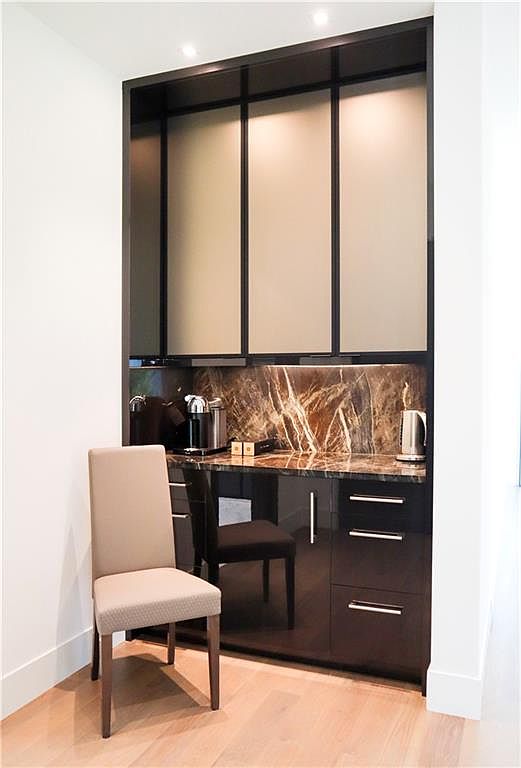 This image showcases a sophisticated built-in coffee bar or beverage station integrated into a wall niche. The space features dark, high-gloss cabinetry, a striking stone backsplash with dramatic veining, and frosted glass upper cabinet doors. A light-toned upholstered chair sits in the foreground, providing a contrast to the sleek, dark finishes of the cabinetry and the light wood flooring.