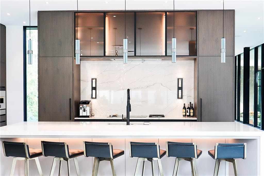 This modern kitchen features a large central island with seating for six, topped with a white stone surface and illuminated by under-counter lighting. The backdrop showcases dark wood cabinetry flanking a white stone backsplash with subtle veining, accented by sleek pendant lights and wall-mounted fixtures. The composition is symmetrical and clean, emphasizing a high-end, contemporary aesthetic suitable for luxury living.