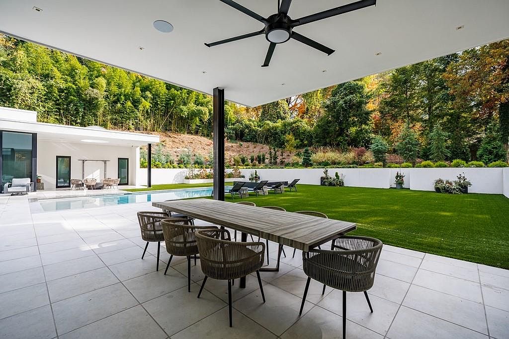 This expansive covered patio features a sleek, modern design with large light-colored floor tiles and a prominent black ceiling fan. A long dining table with woven chairs sits under the overhang, providing a seamless transition to the adjacent swimming pool and manicured lawn. The backyard is framed by a white perimeter wall and a lush, wooded hillside, creating a private and serene outdoor living space.