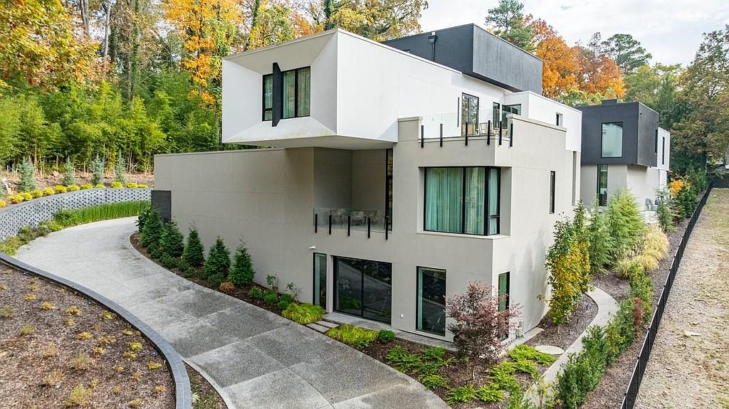 This striking modern residence features a bold, multi-level architectural design with a prominent cantilevered white upper volume and contrasting dark grey accents. The exterior showcases a mix of smooth stucco surfaces, large windows, and a private balcony, all set against a backdrop of mature trees in autumn foliage. A curved concrete driveway leads toward the structure, complemented by manicured landscaping and a decorative retaining wall.