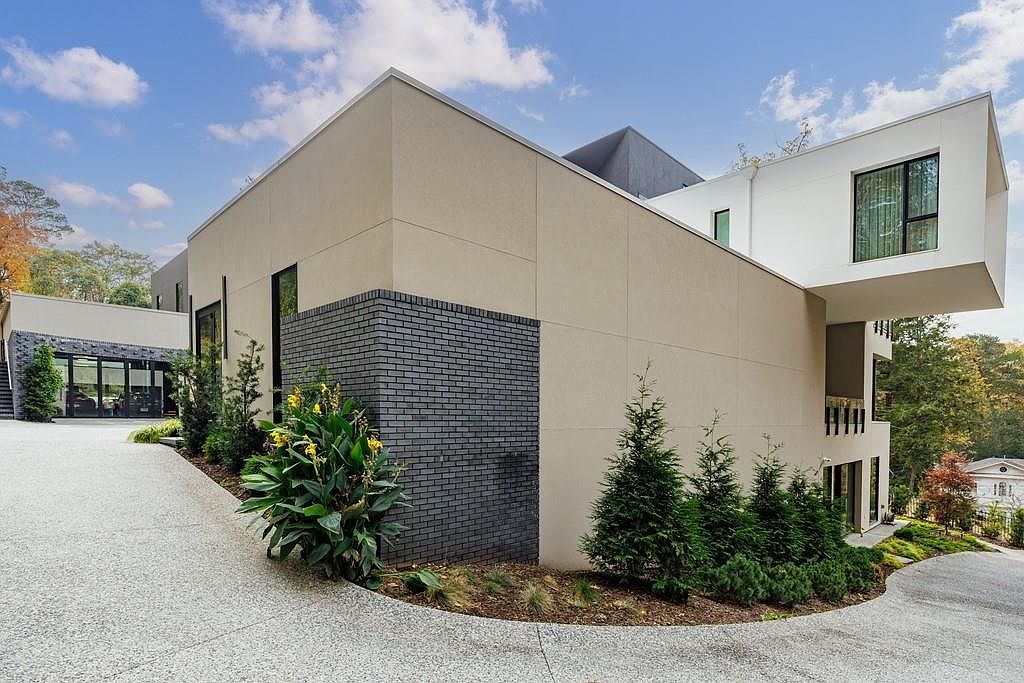 This modern architectural residence features a striking contrast between smooth, light-toned stucco walls and a dark, textured brick section. The design incorporates a cantilevered white upper level that extends over the driveway, creating a bold geometric silhouette. A paved driveway curves around the landscaped front yard, which is accented with young evergreen trees and flowering shrubs under a bright, partly cloudy sky.