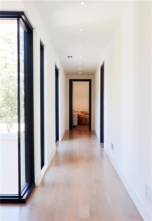 This minimalist hallway features light wood flooring and crisp white walls, creating a bright and airy atmosphere. Tall, black-framed windows line the left side, providing natural light and a view of the outdoors, while recessed ceiling lights guide the way toward a distant bedroom. The clean lines and neutral color palette emphasize a modern, sophisticated architectural style.