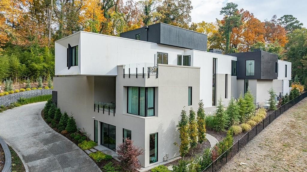 This modern, multi-level residence features a striking architectural design with a mix of white and dark grey exterior finishes. The structure incorporates cantilevered sections, large windows, and a glass-railed balcony, set against a backdrop of mature trees with autumn foliage. A paved driveway leads toward the side of the home, which is bordered by a manicured lawn and a black metal fence.