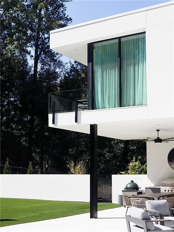 This modern architectural home features a striking white cantilevered upper level with floor-to-ceiling glass sliding doors opening onto a glass-railed balcony. Below, a covered patio area provides an outdoor living space complete with a grill, dining set, and a ceiling fan, overlooking a manicured lawn. The clean lines and minimalist aesthetic are complemented by a dark structural support column and a backdrop of mature trees.