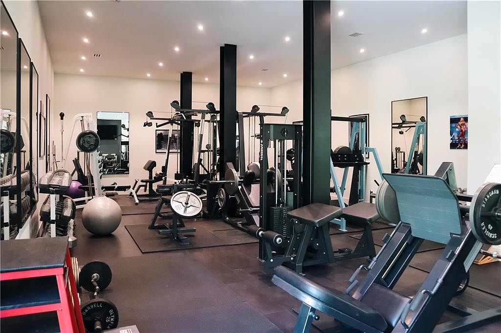 This spacious home gym features a variety of professional-grade strength training equipment, including a leg press machine, cable stations, and a rack of dumbbells. The room is characterized by its dark, industrial-style support columns, mirrored walls that enhance the sense of space, and recessed ceiling lighting. The dark rubberized flooring provides a functional foundation for this dedicated fitness area.