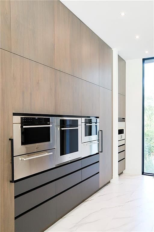 This modern kitchen features a sleek, minimalist wall of light wood cabinetry integrated with a suite of stainless steel built-in appliances. Below the appliances, a row of dark grey drawers provides a striking contrast, while the floor is finished in a light-colored, veined stone tile. A tall window at the end of the hallway allows natural light to flood the space, emphasizing the clean lines and contemporary aesthetic.