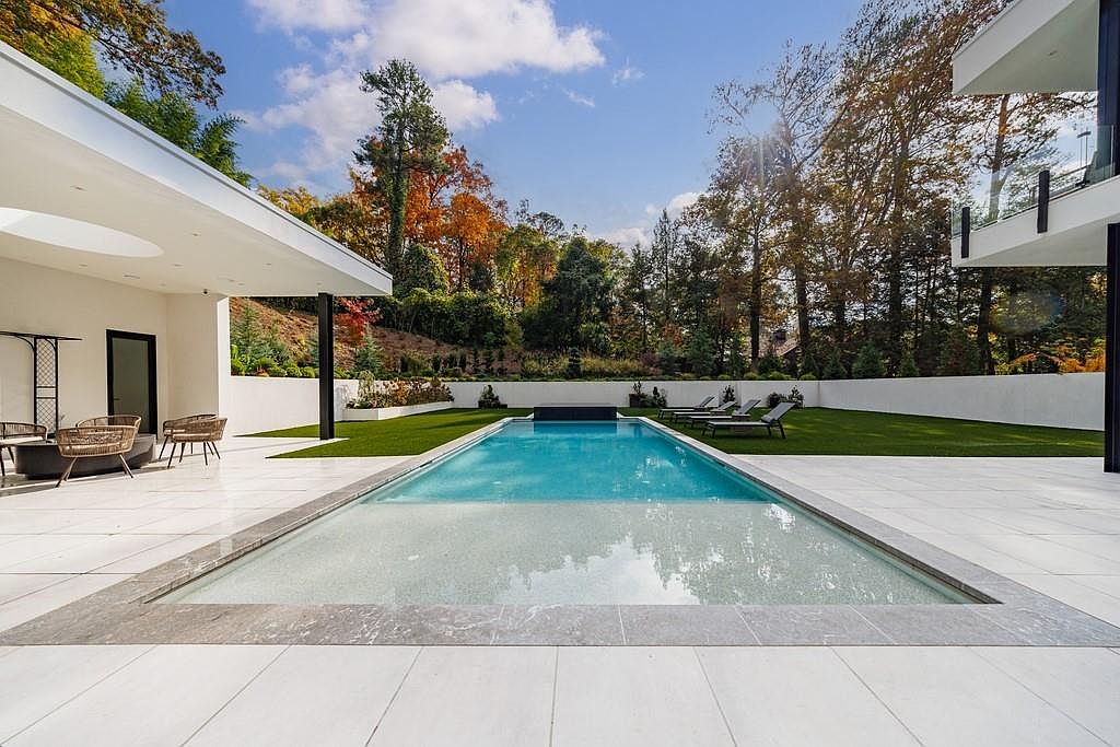 This modern backyard features a sleek, rectangular swimming pool as the central focal point, surrounded by light-toned stone decking. To the left, a covered patio area provides a shaded lounge space, while a manicured lawn with several sun loungers extends toward the rear wall. The scene is framed by mature trees with autumn foliage, creating a serene and luxurious outdoor living environment.