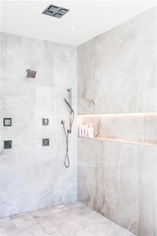 This luxurious walk-in shower features large-format, light-toned stone wall tiles with subtle veining and a matching tiled floor. The space is equipped with a modern rainfall showerhead, a handheld sprayer, and multiple wall-mounted body jets, all finished in a sleek metallic tone. A recessed, illuminated niche provides convenient storage for toiletries, adding a sophisticated and functional touch to the contemporary design.