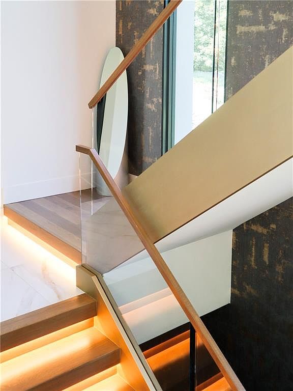 This modern staircase features floating wood treads illuminated by integrated under-step lighting, creating a warm and inviting atmosphere. A glass railing with a sleek wood handrail provides a clean, contemporary aesthetic, while a textured, dark-toned wall covering adds depth and sophistication to the space. The perspective captures the elegant transition between levels, highlighting the high-end finishes and architectural detail.