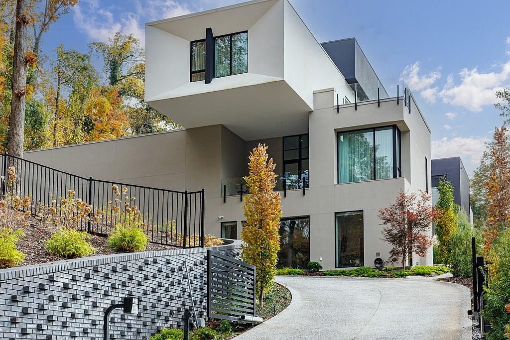 This modern architectural residence features a striking cantilevered upper level with a clean, minimalist aesthetic. The exterior combines smooth light-toned surfaces with dark accents, complemented by a textured brick retaining wall and a paved driveway leading toward the entrance. Large windows and glass railings enhance the contemporary design, set against a backdrop of mature trees and a landscaped hillside.