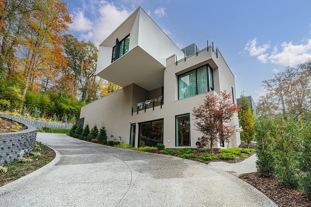 This modern architectural home features a striking cantilevered upper level that creates a dramatic overhang above the driveway. The exterior is finished in a smooth, neutral-toned stucco, complemented by large windows and a glass-railed balcony. A curved stone retaining wall lines the driveway, while the surrounding landscape includes mature trees and manicured shrubbery, emphasizing a sophisticated, contemporary aesthetic.