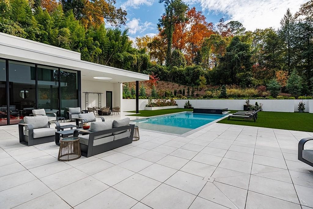 This modern outdoor living space features a rectangular swimming pool set within a large, light-colored stone patio. The area includes a comfortable seating arrangement with gray cushioned furniture and a separate lounge area with sun loungers on a manicured lawn. The scene is framed by a contemporary home with large glass walls and a lush, wooded backdrop with vibrant autumn foliage.