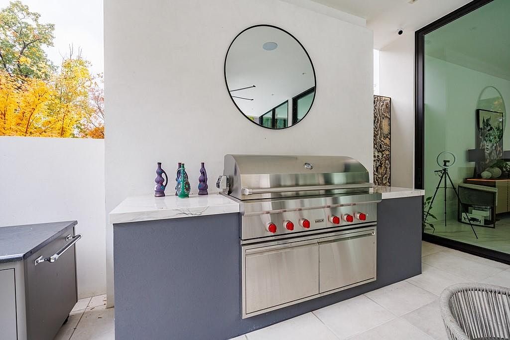 This outdoor kitchen area features a sleek, stainless steel grill set into a dark-toned base with a light-colored stone countertop. A circular mirror hangs on the white wall above the grill, reflecting the surrounding space, while three decorative vases sit to the left. The area is adjacent to a large glass sliding door that provides a view into the interior, creating a seamless transition between indoor and outdoor living.