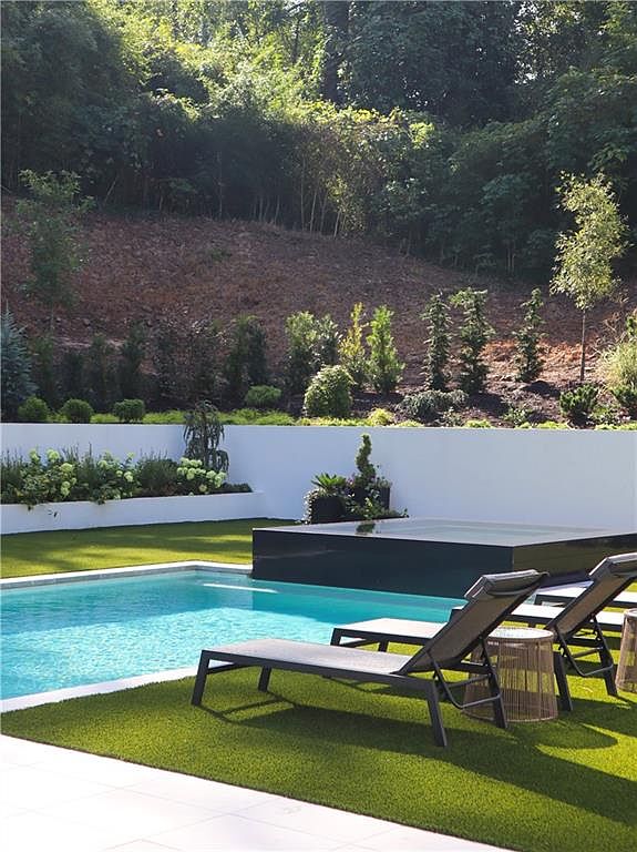 This serene outdoor space features a rectangular swimming pool paired with a sleek, dark-toned raised spa. Two modern lounge chairs are positioned on the manicured artificial turf, facing the water and a clean white retaining wall that separates the pool area from a lush, terraced hillside. The scene offers a tranquil, resort-like atmosphere perfect for relaxation and outdoor entertaining.
