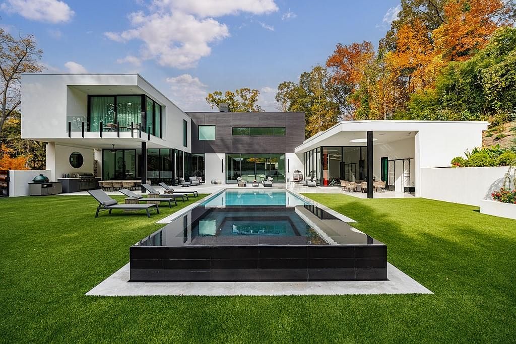 This modern luxury residence features a striking architectural design with a mix of white stucco and dark, textured paneling. The rear of the home opens onto a manicured lawn and a sleek, rectangular swimming pool with an integrated spa. Large glass walls and sliding doors create a seamless transition between the indoor living spaces and the outdoor patio area, which is furnished with lounge chairs and an outdoor kitchen.