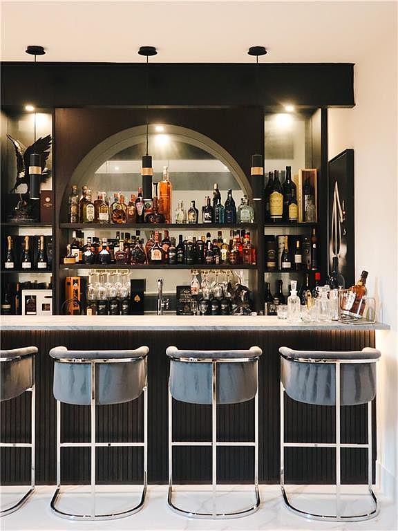 This sophisticated home bar area features a sleek, dark-toned design with a central arched display shelf illuminated by recessed lighting. The bar is equipped with a light-colored stone countertop, a row of modern grey velvet bar stools with metallic frames, and a well-stocked collection of spirits and glassware. The overall aesthetic is upscale and contemporary, creating an inviting space for entertaining.