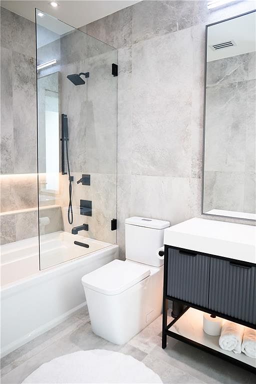 This modern bathroom features a sleek, minimalist aesthetic with large-format grey stone-look wall tiles and a glass-enclosed shower area. The space includes a contemporary white toilet, a floating vanity with a fluted dark grey cabinet, and a clean white countertop. A tall, thin mirror hangs above the vanity, and the overall design emphasizes clean lines and a sophisticated, neutral color palette.