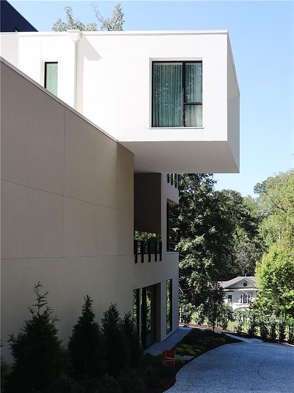 This side view of a modern residence highlights a striking cantilevered upper level that extends over a recessed outdoor space. The structure features a clean, minimalist aesthetic with smooth, light-toned exterior walls and large, rectangular windows. A gravel driveway leads toward the back of the property, where lush green trees and a glimpse of a traditional-style home in the distance create a contrast between contemporary and classic architectural styles.