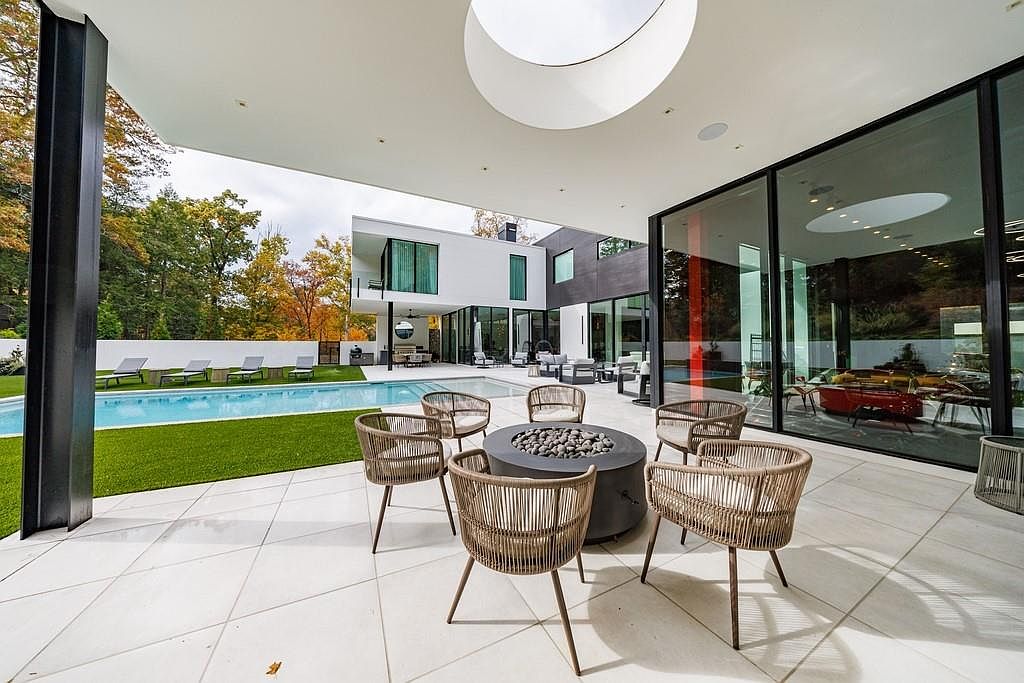 This modern outdoor living space features a covered patio area with a circular fire pit surrounded by woven chairs, set against a backdrop of a sleek, contemporary home. The area overlooks a pristine swimming pool and a manicured lawn, with large glass walls reflecting the surrounding autumn trees. A unique circular skylight is integrated into the patio ceiling, enhancing the architectural interest of this sophisticated, high-end residence.