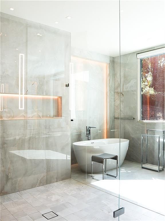 This luxurious primary bathroom features a sleek, freestanding soaking tub positioned in front of a large window that allows natural light to fill the space. The room is finished with large-format stone wall tiles and a matching tiled floor, complemented by a minimalist stool and a chrome towel rack. A glass-enclosed shower area with integrated recessed lighting creates a clean, modern, and spa-like atmosphere.