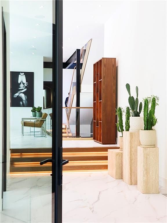 This interior view captures a sophisticated entryway leading to a staircase, framed by a sleek black-trimmed glass door. The space features polished light-toned flooring with subtle veining, a tall wooden shelving unit, and a series of stone pedestals topped with potted cacti. A glimpse into the adjacent room reveals a minimalist seating area and a large black-and-white art piece, creating a modern and gallery-like atmosphere.
