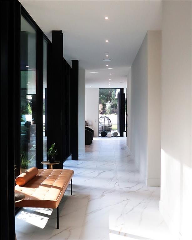 This sleek, modern hallway features polished white stone flooring with subtle veining and a long, tan leather bench positioned against a wall of black-framed glass panels. The perspective looks down a bright, linear corridor toward a distant doorway that opens to an outdoor space, creating a sense of depth and openness. Recessed ceiling lighting provides a clean, minimalist aesthetic that emphasizes the home's contemporary architectural design.