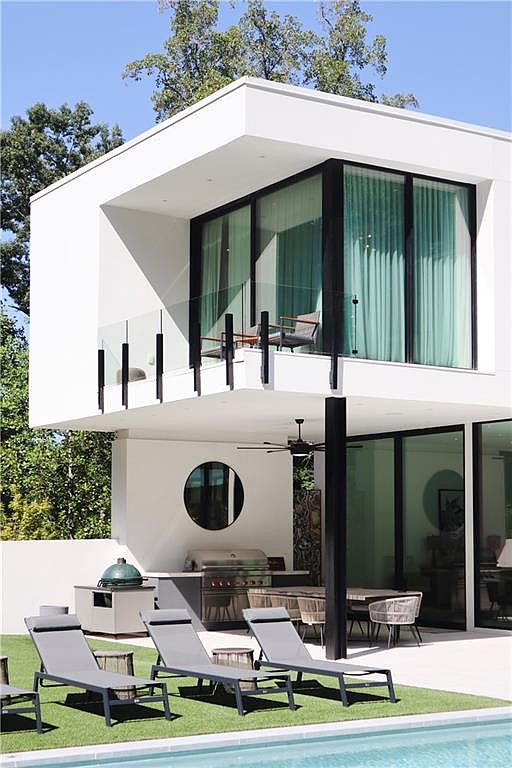 This modern, minimalist residence features a striking cantilevered upper balcony with glass railings and floor-to-ceiling windows. The ground level offers a seamless indoor-outdoor transition to a covered patio area equipped with an outdoor kitchen, grill, and dining space, all overlooking a pristine swimming pool. The clean white architectural lines and contemporary lounge furniture create a sophisticated, resort-like atmosphere perfect for luxury outdoor living.