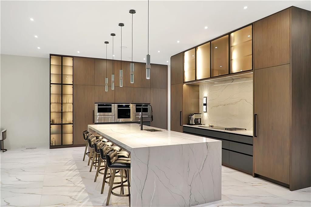 This sophisticated, modern kitchen features a large central island with a stone waterfall edge and integrated seating for four. The space is defined by dark wood cabinetry, illuminated glass-front upper cabinets, and a sleek stone backsplash. A series of minimalist pendant lights hangs above the island, creating a clean, high-end aesthetic against the light-toned stone flooring.