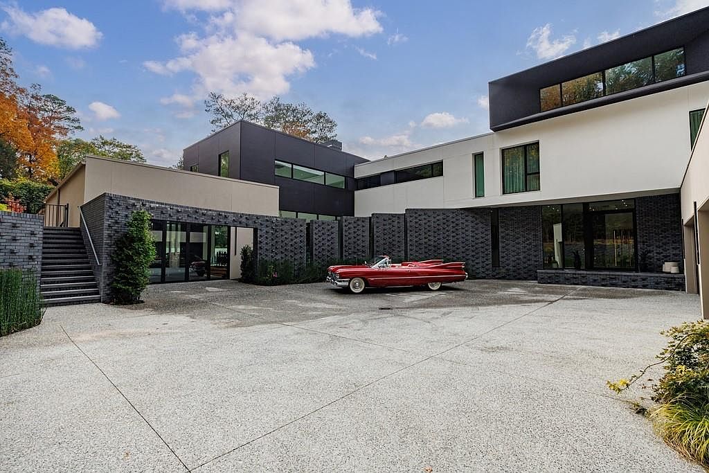 This striking modern residence features a multi-level architectural design with a mix of dark charcoal brick and smooth white exterior panels. A vibrant red vintage convertible is parked in the center of the expansive, light-colored concrete motor court, which is framed by a unique textured brick wall. The composition captures the home from a low-angle perspective, emphasizing the clean lines and the contrast between the dark masonry and the bright, contemporary structure.