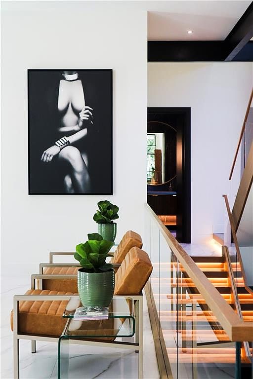 This sophisticated hallway features a modern staircase with illuminated wooden treads and a sleek glass railing. Two tan leather chairs with metal frames are positioned alongside a glass side table, each holding a potted plant, creating a stylish seating area beneath a large black-and-white art piece. The space is defined by clean white walls, polished light-colored flooring, and a dark-framed doorway leading to an adjacent room.