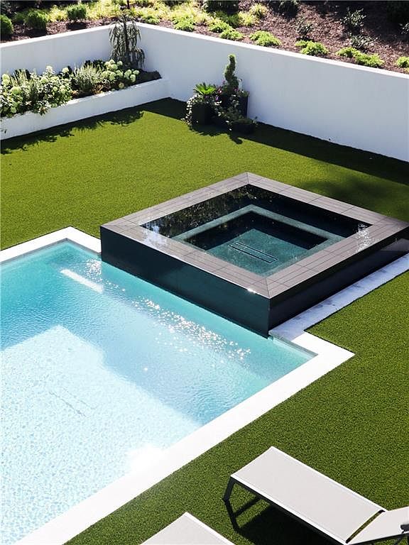 This high-angle aerial view captures a modern backyard oasis featuring a rectangular swimming pool adjacent to a raised, dark-tiled square spa. The area is surrounded by vibrant green artificial turf and a clean white perimeter wall, creating a sharp, minimalist aesthetic. A portion of a lounge chair is visible in the foreground, emphasizing the space as a private retreat for relaxation.