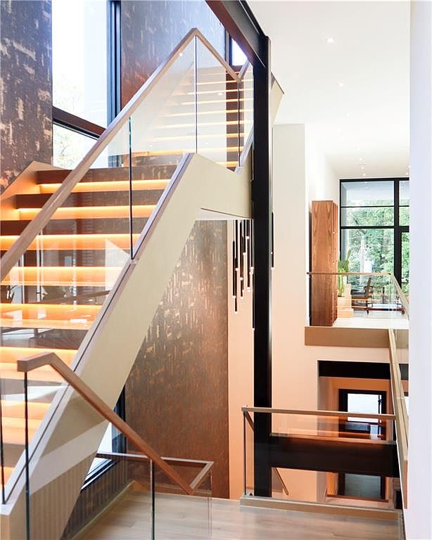 This striking interior view captures a modern staircase featuring illuminated treads and glass railings, set against a textured, dark-toned wall. The perspective looks down from a landing, showcasing the open, multi-level architectural design and a vertical light fixture hanging in the stairwell. Large windows at the end of the hallway provide natural light and a view of the surrounding greenery, emphasizing the home's contemporary and airy aesthetic.