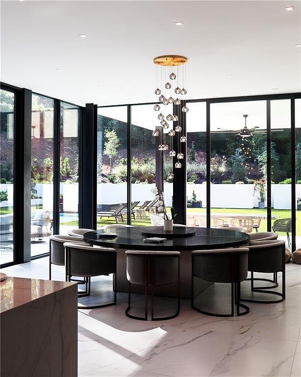 This sophisticated dining room features a large, circular black table surrounded by modern, curved-back chairs, positioned centrally under a striking multi-pendant light fixture. The space is defined by expansive floor-to-ceiling windows that provide an unobstructed view of the lush backyard and swimming pool area. The room is finished with polished, light-colored stone flooring, creating a seamless and airy atmosphere that emphasizes the indoor-outdoor connection.