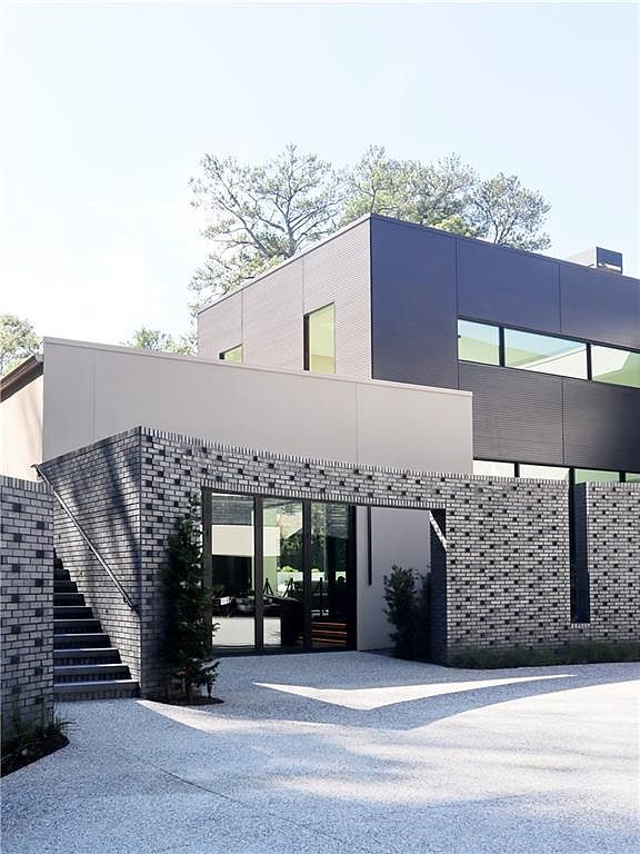 This modern architectural entryway features a striking combination of dark, textured brickwork and smooth, light-toned panels. A set of stairs leads up to a glass-fronted entrance, while the geometric design and clean lines create a sophisticated, contemporary aesthetic. The perspective captures the home's unique facade and the gravel driveway, emphasizing its bold, minimalist curb appeal.