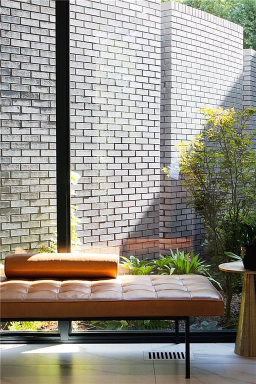 This interior space features a sleek, tan leather bench with a matching bolster pillow positioned in front of large, floor-to-ceiling windows. The windows look out onto a private garden area with lush greenery and a textured, light-colored brick wall. A small, metallic side table sits to the right, and the room is finished with light-toned flooring, creating a serene and minimalist atmosphere.