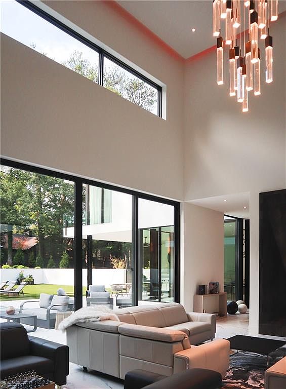This expansive, double-height living room features a contemporary design with a neutral color palette and a striking modern chandelier composed of vertical glass rods. Large, multi-panel sliding glass doors provide a seamless transition to the outdoor patio and garden, flooding the space with natural light. The room is furnished with a modular light-toned sofa and a dark accent chair, creating a sophisticated and airy atmosphere.