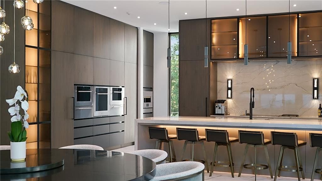 This sophisticated, modern kitchen features dark wood cabinetry, a large central island with bar seating, and integrated stainless steel appliances. The space is illuminated by elegant pendant lights and under-cabinet lighting, highlighting the stone backsplash and sleek countertops. A dining area with a round table and a white orchid centerpiece is visible in the foreground, creating a seamless and luxurious open-concept living space.