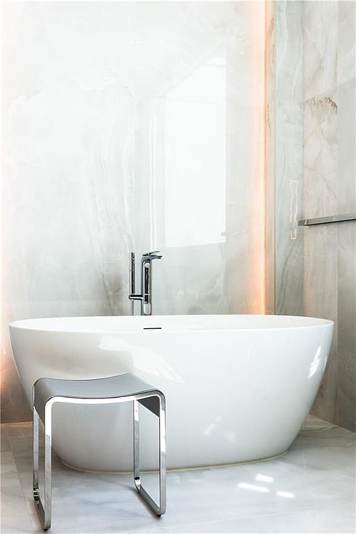 This elegant bathroom features a sleek, white freestanding soaking tub positioned against a wall clad in large-format, light-toned stone panels with subtle veining. A modern, polished chrome faucet rises from the deck of the tub, complemented by a minimalist, metallic stool placed in the foreground. The space is illuminated by soft, warm accent lighting integrated into the wall, creating a serene and luxurious spa-like atmosphere.