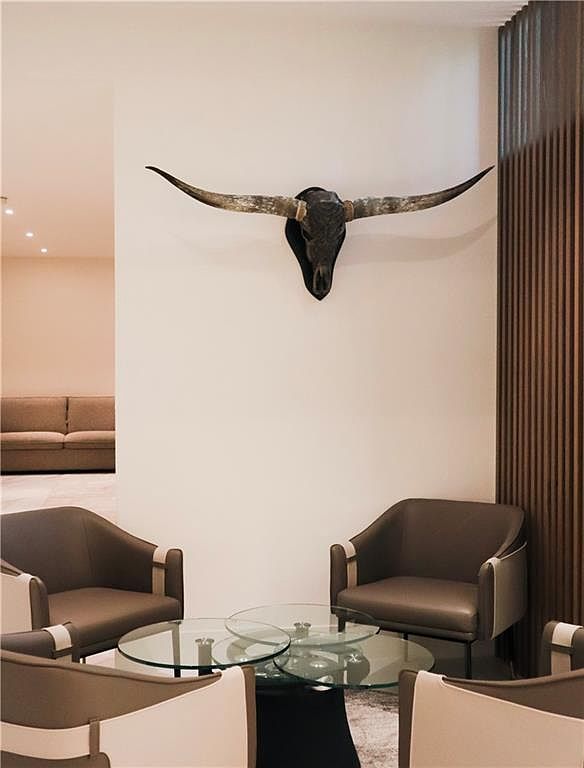 This sophisticated seating area features a pair of modern, curved armchairs upholstered in neutral tones, arranged around a tiered glass coffee table. A striking longhorn skull wall mount serves as a bold focal point above the seating arrangement, set against a clean, minimalist wall. To the right, a vertical wood-slat wall adds texture and warmth, while a glimpse of a sofa in the background suggests an open-concept layout.