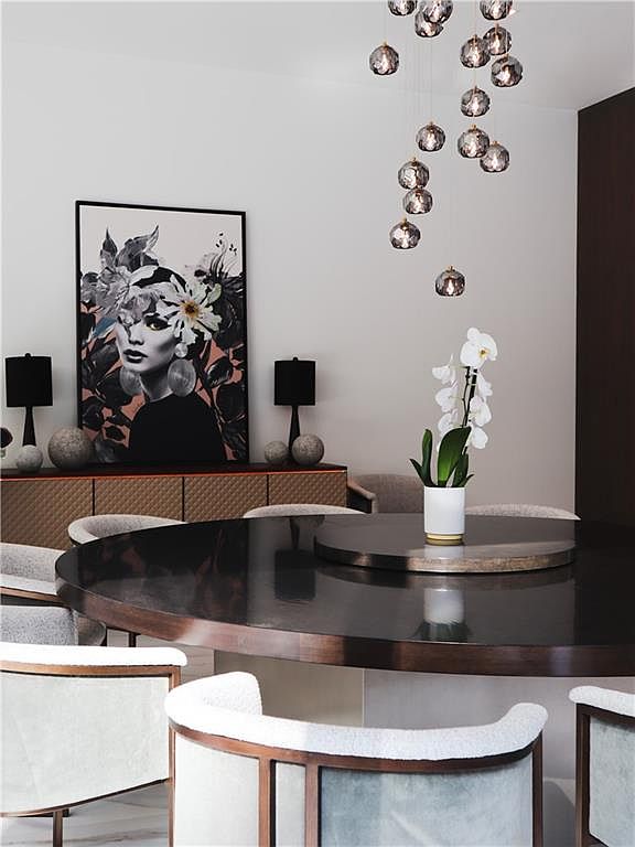 This sophisticated dining area features a large, dark circular table surrounded by modern chairs with light-colored, textured upholstery. A striking piece of wall art depicting a stylized portrait serves as a focal point above a long, textured sideboard, while a cluster of elegant, spherical pendant lights hangs above the table. The room is accented by a white orchid in a simple pot, creating a refined and contemporary atmosphere.