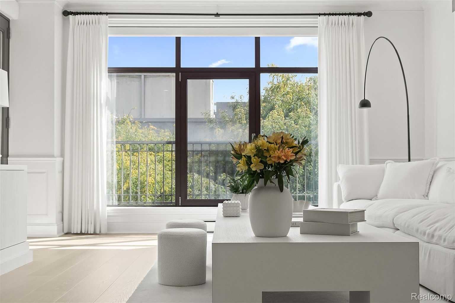 This is a bright and airy living room featuring a large window with a view of greenery and a blue sky. The room is decorated in a minimalist style with white walls, light wood floors, and neutral-toned furniture. A modern arc lamp and a vase of flowers on the coffee table add subtle accents to the space.